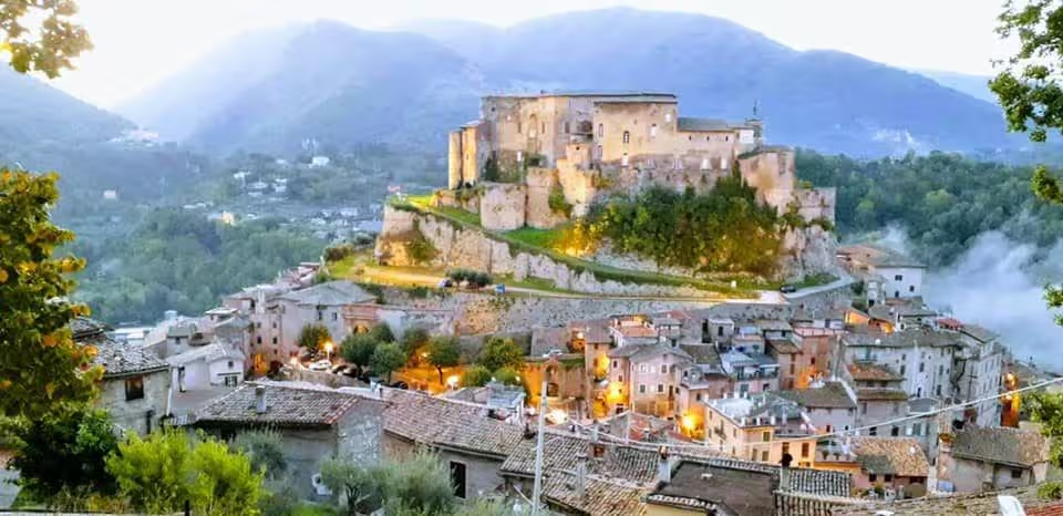 Veduta panoramica del borgo medievale di Subiaco al crepuscolo: il paese terrazzato sulla collina con la Rocca Abbaziale in cima, i tetti in coppi e la nebbia nella valle