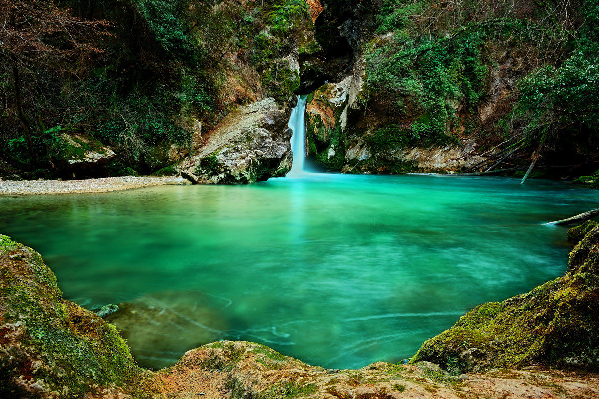 Polla d'acqua smeraldo del Lago di San Benedetto a Subiaco con cascata setosa, massi coperti di musco e vegetazione selvatica