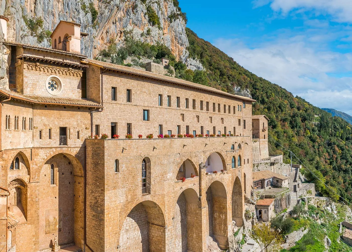 Il Sacro Speco di Subiaco aggrappato alla parete rocciosa, con la terrazza di fiori rossi e la foresta verde della valle dell'Aniene
